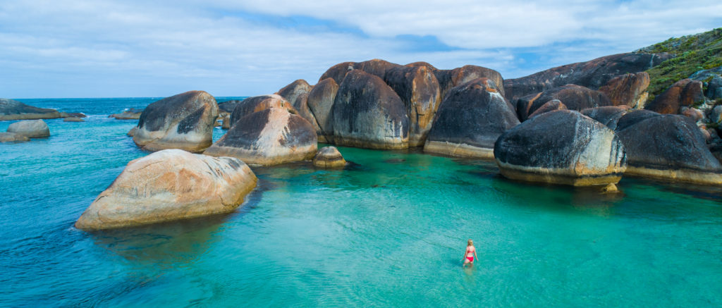 landscape image of girl in peppermint coloured water with boulders to show epic beaches en route along The South West Edge